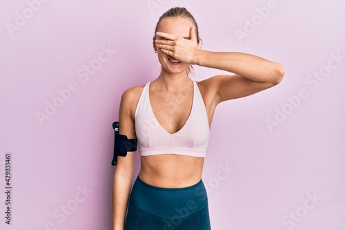 Beautiful blonde woman wearing sportswear and arm band smiling and laughing with hand on face covering eyes for surprise. blind concept. © Krakenimages.com