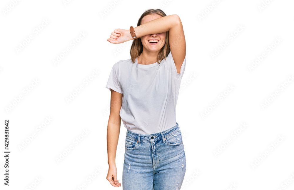 Fototapeta premium Beautiful young blonde woman wearing casual white t shirt covering eyes with arm smiling cheerful and funny. blind concept.