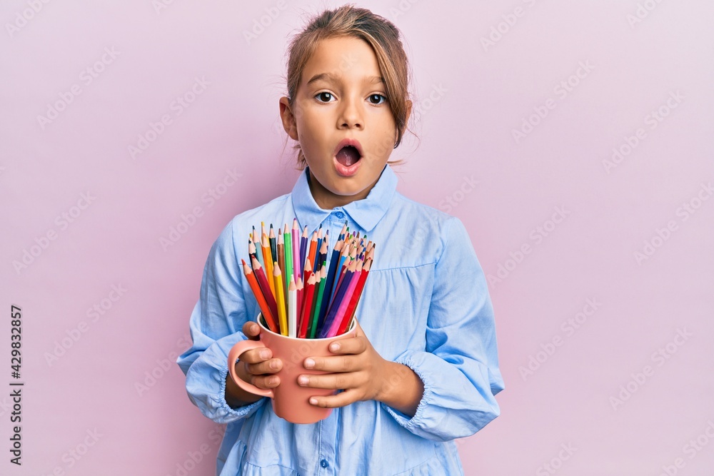 Little beautiful girl holding colored pencils afraid and shocked with surprise and amazed expression, fear and excited face. Little beautiful girl holding colored pencils afraid and shocked with surprise and amazed expression, fear and excited face.