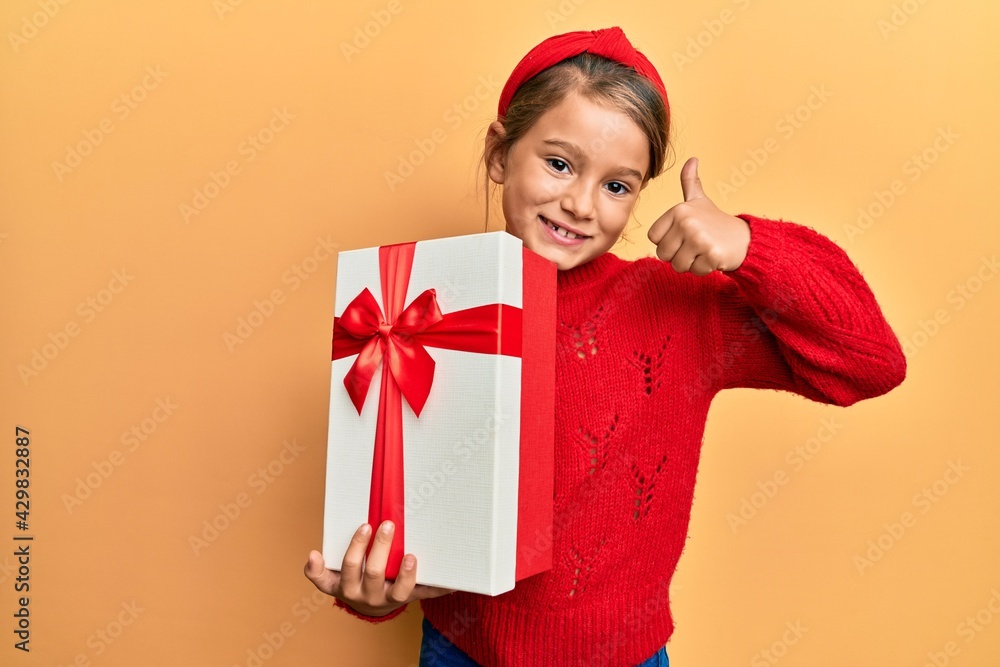 Little beautiful girl holding gift smiling happy and positive, thumb up doing excellent and approval sign Little beautiful girl holding gift smiling happy and positive, thumb up doing excellent and approval sign
