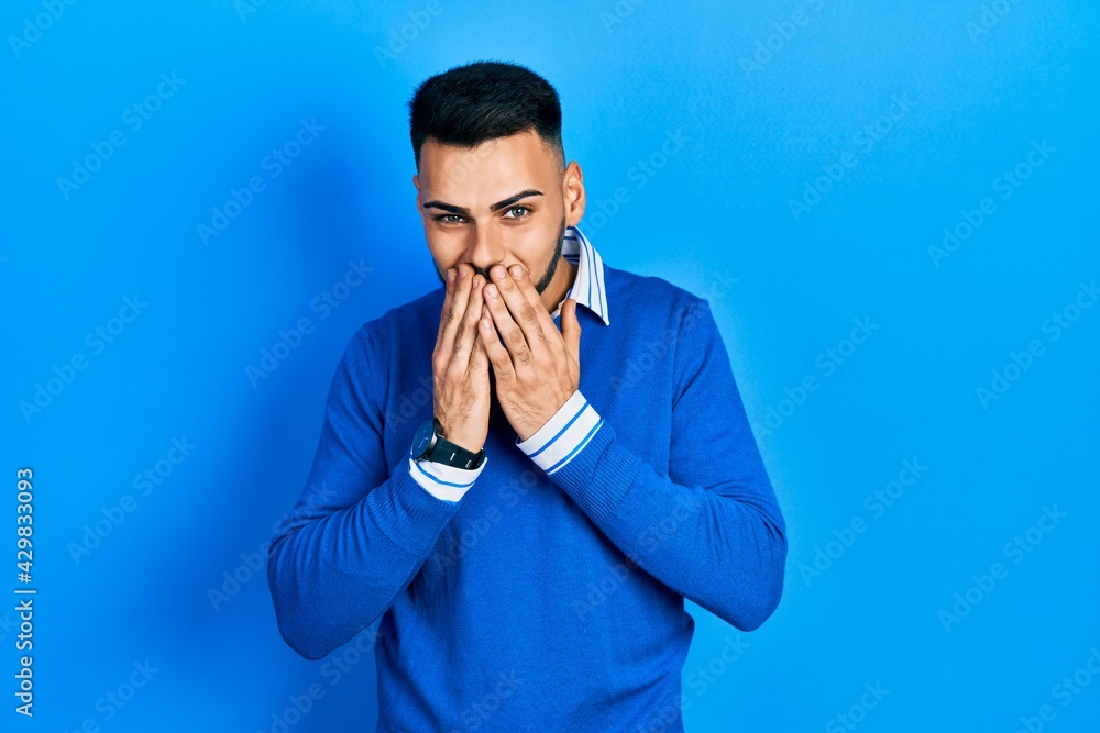 Young hispanic man with beard wearing casual blue sweater laughing and embarrassed giggle covering mouth with hands, gossip and scandal concept