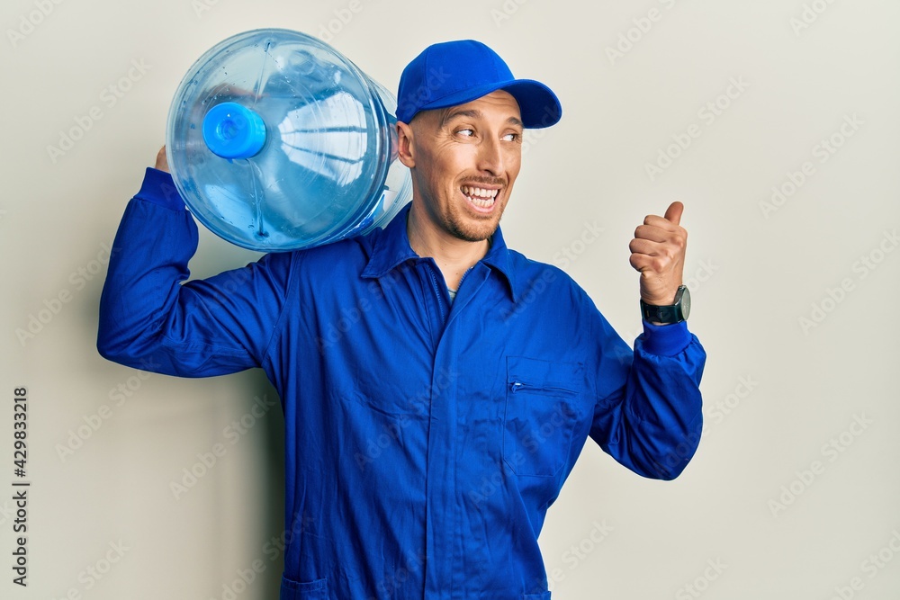 Bald courier man with beard holding a gallon bottle of water for delivery pointing thumb up to the side smiling happy with open mouth