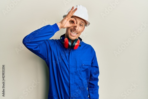 Bald man with beard wearing builder jumpsuit uniform and hardhat smiling happy doing ok sign with hand on eye looking through fingers