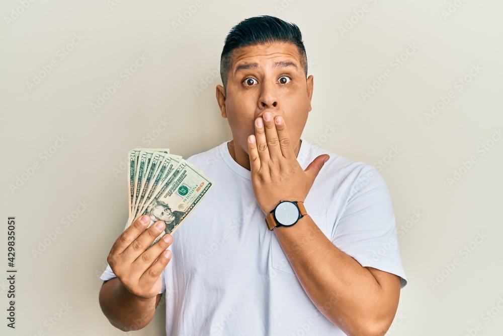 Young latin man holding 20 dollars banknotes covering mouth with hand, shocked and afraid for mistake. surprised expression Young latin man holding 20 dollars banknotes covering mouth with hand, shocked and afraid for mistake. surprised expression