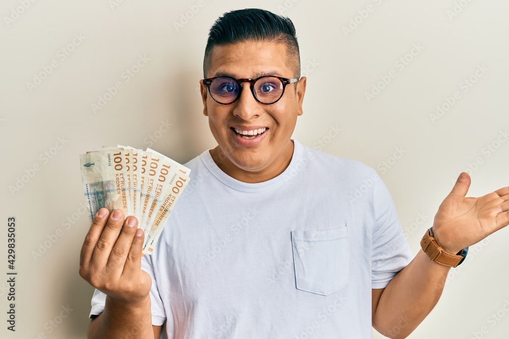 Young latin man holding 100 danish krone banknotes celebrating achievement with happy smile and winner expression with raised hand Young latin man holding 100 danish krone banknotes celebrating achievement with happy smile and winner expression with raised hand