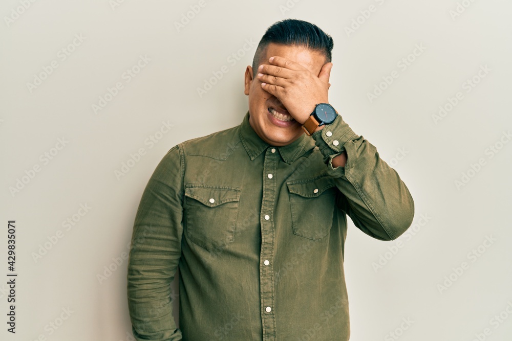 Young latin man wearing casual clothes smiling and laughing with hand on face covering eyes for surprise. blind concept. Young latin man wearing casual clothes smiling and laughing with hand on face covering eyes for surprise. blind concept.