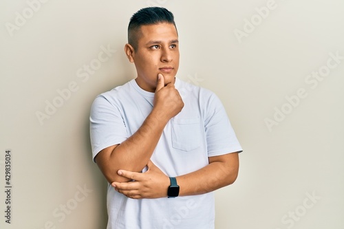 Young latin man wearing casual white t shirt with hand on chin thinking about question, pensive expression. smiling with thoughtful face. doubt concept.