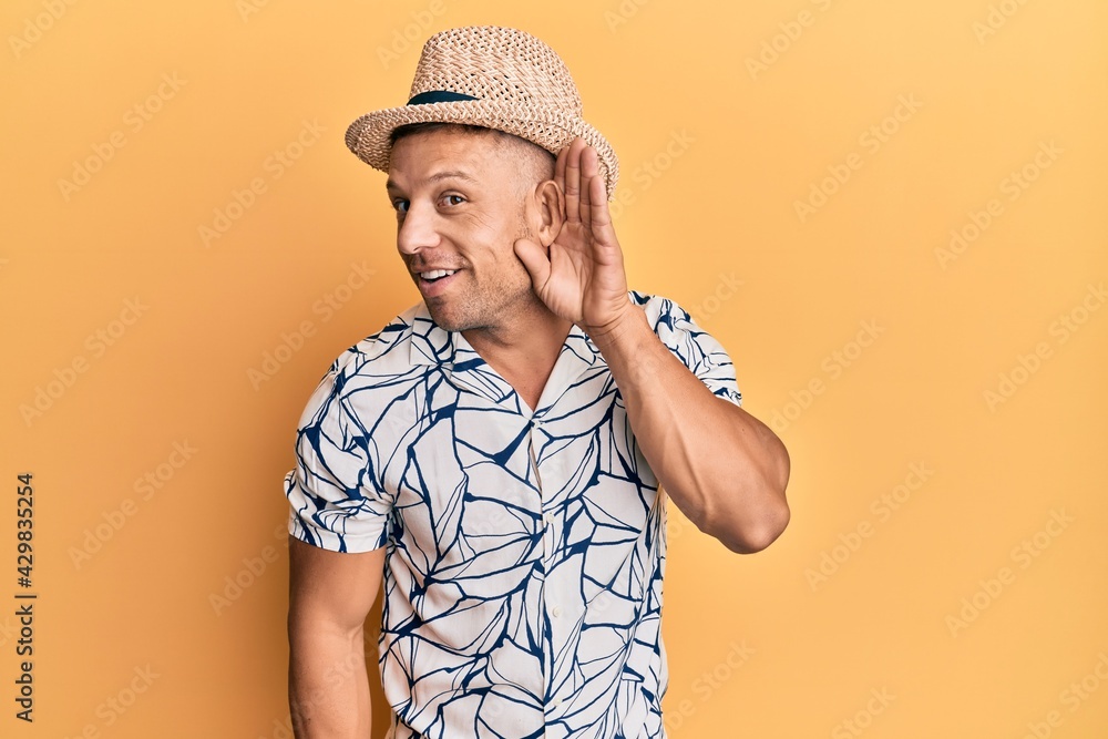 Handsome muscle man wearing summer hat smiling with hand over ear listening and hearing to rumor or gossip. deafness concept. Handsome muscle man wearing summer hat smiling with hand over ear listening and hearing to rumor or gossip. deafness concept.