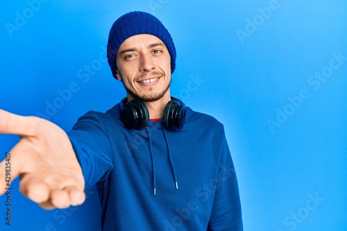 Hispanic young man wearing sweatshirt and headphones smiling friendly offering handshake as greeting and welcoming. successful business.