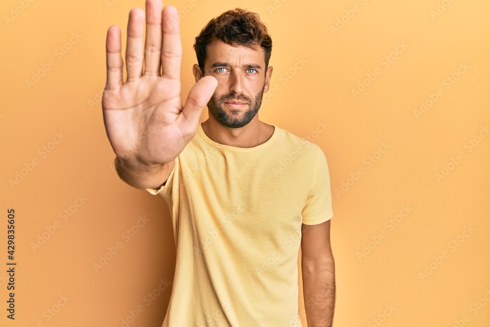 Handsome man with beard wearing casual yellow tshirt over yellow background doing stop sing with palm of the hand. warning expression with negative and serious gesture on the face. Handsome man with beard wearing casual yellow tshirt over yellow background doing stop sing with palm of the hand. warning expression with negative and serious gesture on the face.