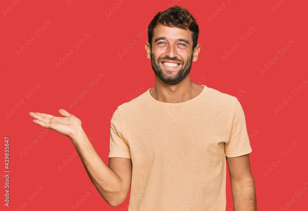 Handsome young man with beard wearing casual tshirt smiling cheerful presenting and pointing with palm of hand looking at the camera. Handsome young man with beard wearing casual tshirt smiling cheerful presenting and pointing with palm of hand looking at the camera.