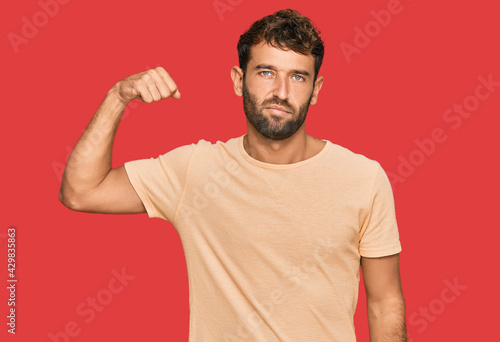 Handsome young man with beard wearing casual tshirt strong person showing arm muscle, confident and proud of power