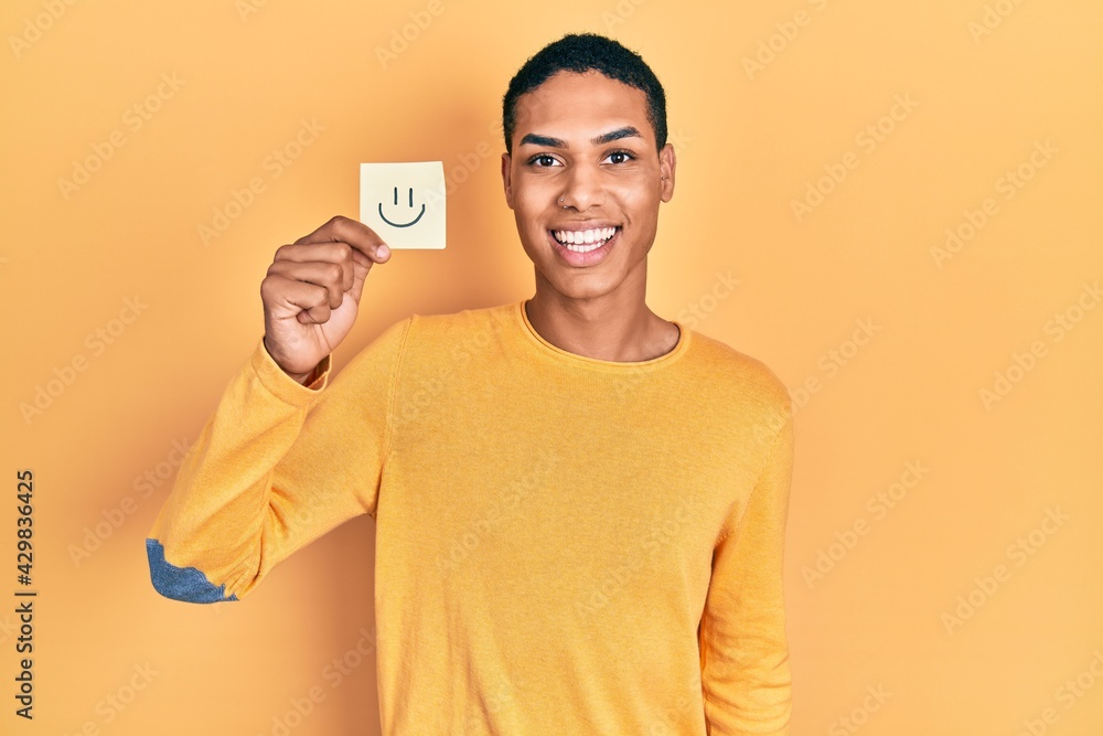 Young african american guy holding smile reminder looking positive and happy standing and smiling with a confident smile showing teeth Young african american guy holding smile reminder looking positive and happy standing and smiling with a confident smile showing teeth