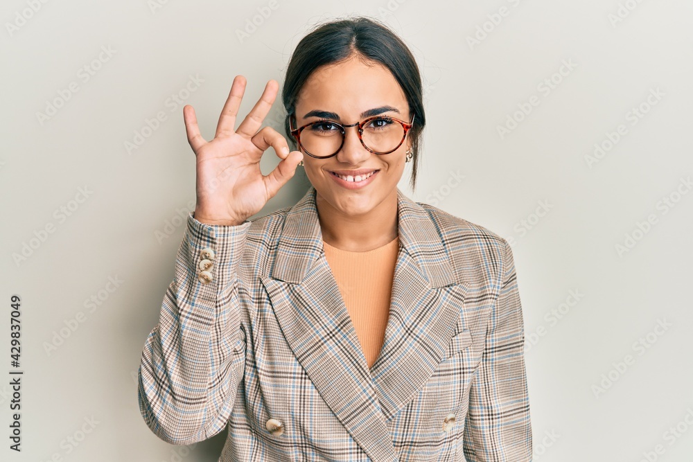 Young brunette woman wearing business jacket and glasses smiling positive doing ok sign with hand and fingers. successful expression. Young brunette woman wearing business jacket and glasses smiling positive doing ok sign with hand and fingers. successful expression.