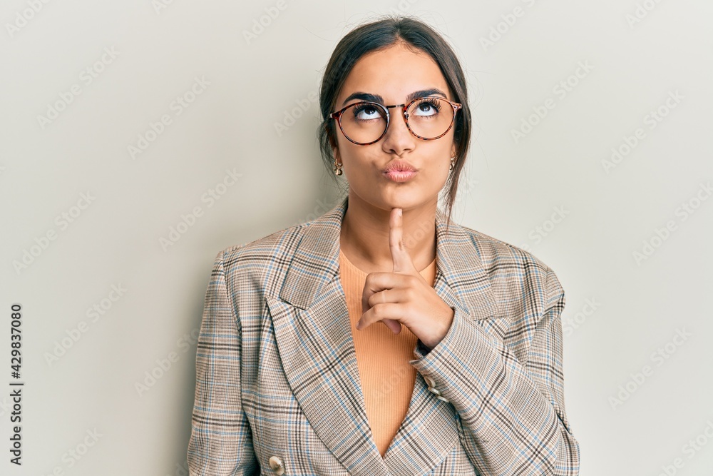 Young brunette woman wearing business jacket and glasses thinking concentrated about doubt with finger on chin and looking up wondering