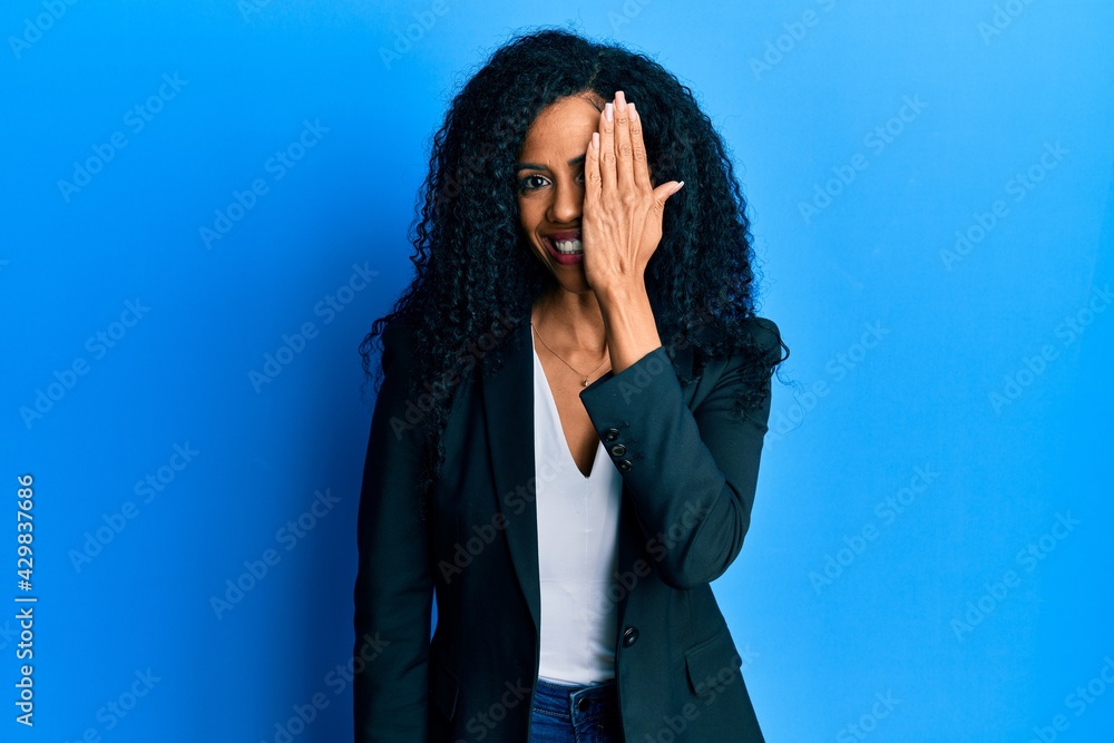 Middle age african american woman wearing business clothes covering one eye with hand, confident smile on face and surprise emotion. Middle age african american woman wearing business clothes covering one eye with hand, confident smile on face and surprise emotion.