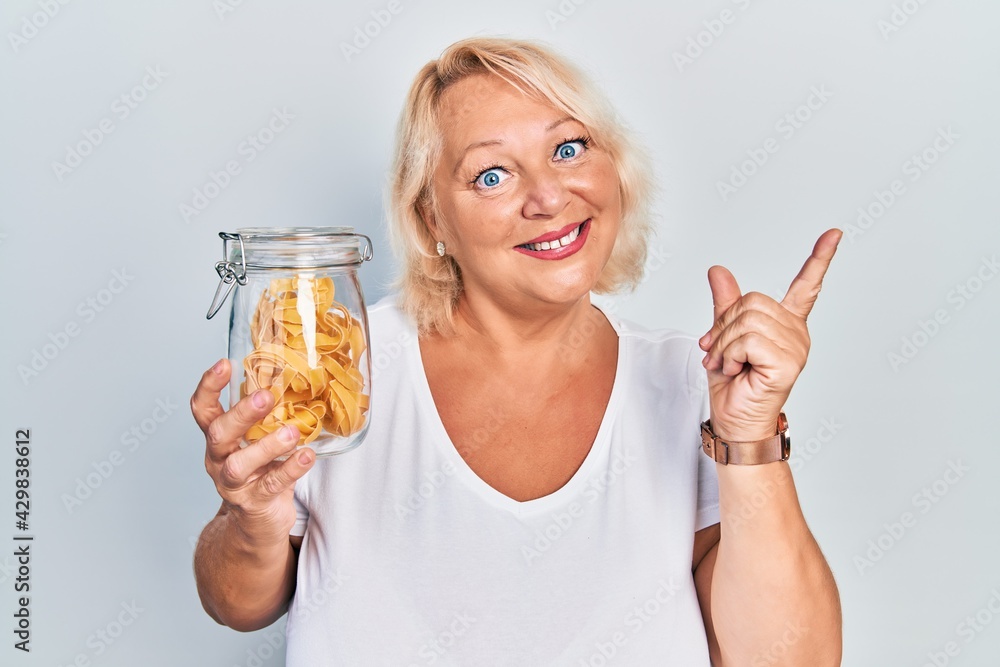 Middle age blonde woman holding uncooked noodles smiling happy pointing with hand and finger to the side Middle age blonde woman holding uncooked noodles smiling happy pointing with hand and finger to the side