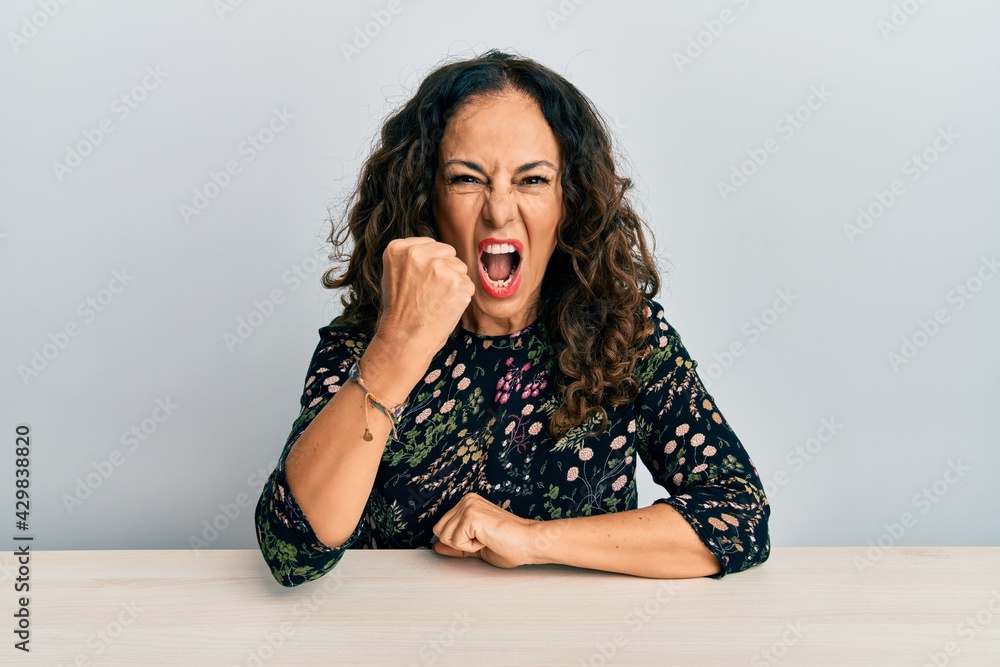 Beautiful middle age woman wearing casual clothes sitting on the table angry and mad raising fist frustrated and furious while shouting with anger. rage and aggressive concept. Beautiful middle age woman wearing casual clothes sitting on the table angry and mad raising fist frustrated and furious while shouting with anger. rage and aggressive concept.