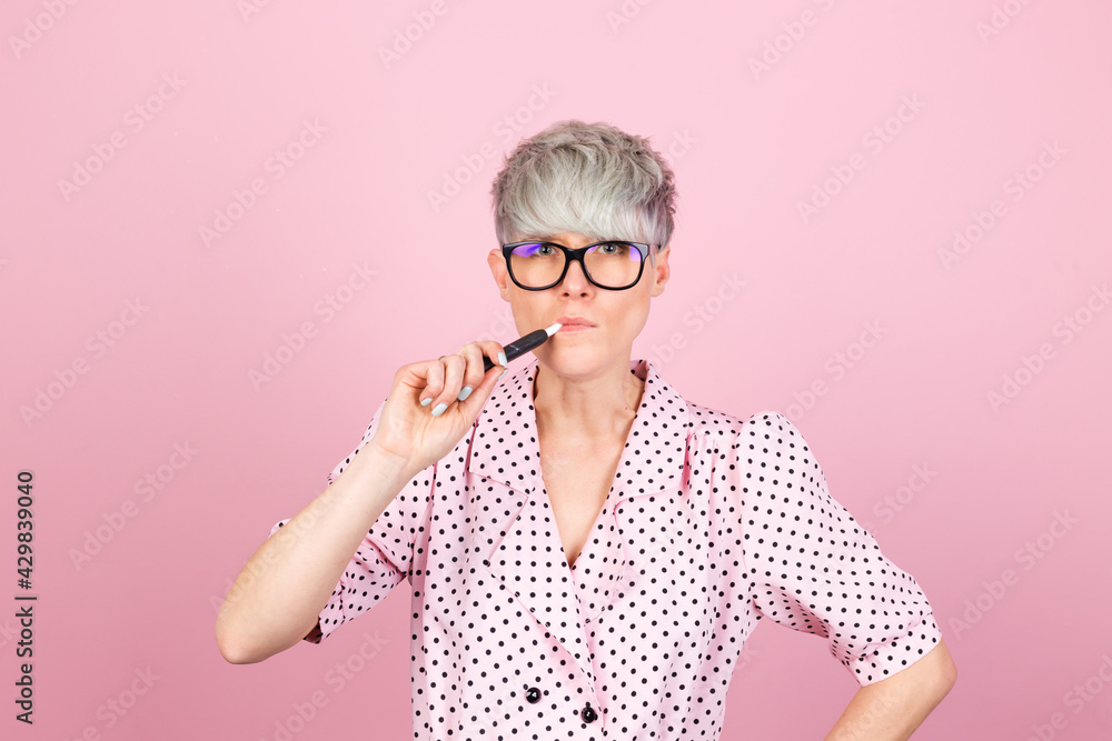 Stylish woman in dress and glasses on pink background holding heated tobacco IQOS system smoking Stylish woman in dress and glasses on pink background holding heated tobacco IQOS system smoking
