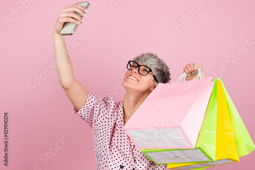 Stylish woman in dress and glasses on pink background holding shopping bags taking selfie on mobile phone happy emotions