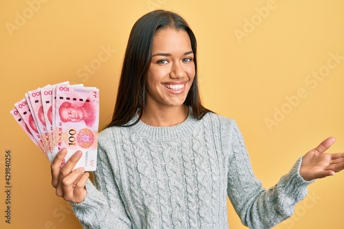 Beautiful hispanic woman holding 100 yuan chinese banknotes celebrating achievement with happy smile and winner expression with raised hand