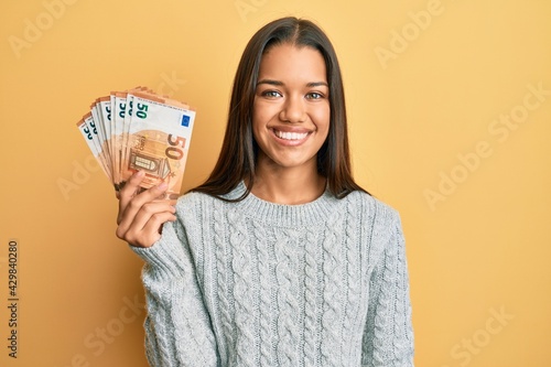 Beautiful hispanic woman holding bunch of 50 euro banknotes looking positive and happy standing and smiling with a confident smile showing teeth