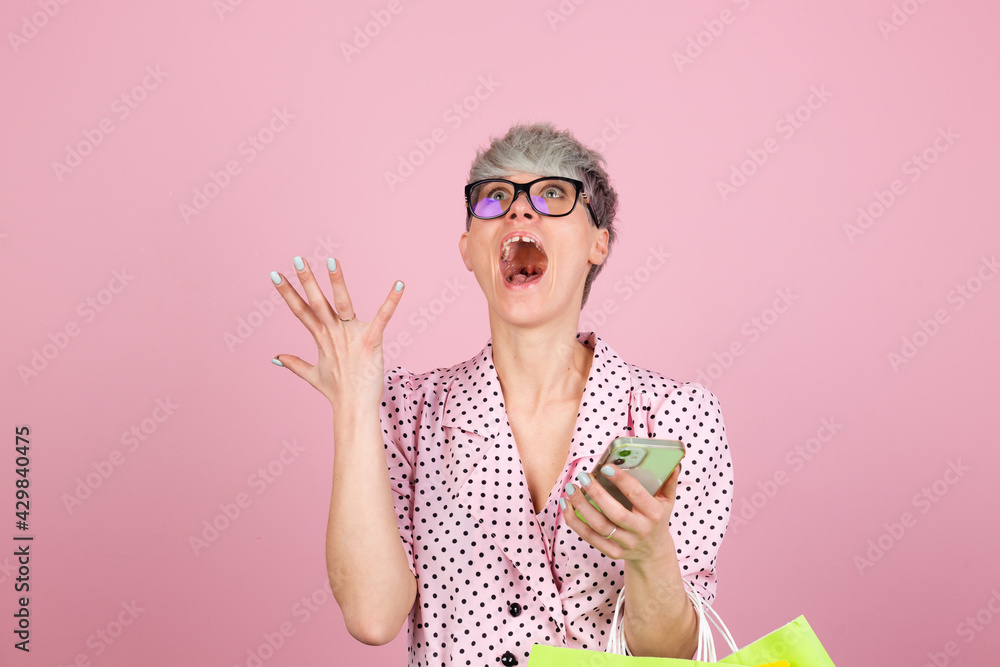 Stylish woman in dress and glasses on pink background holding shopping bags looking searching on mobile phone happy shocked excited amazed