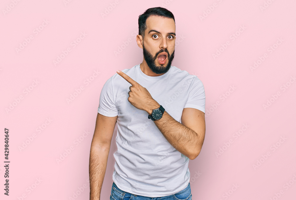 Young man with beard wearing casual white t shirt surprised pointing with finger to the side, open mouth amazed expression. Young man with beard wearing casual white t shirt surprised pointing with finger to the side, open mouth amazed expression.