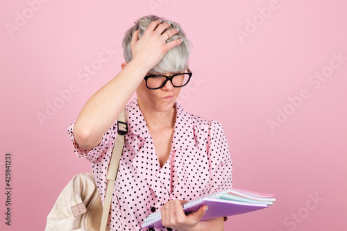 Stylish woman in dress and glasses on pink background with notebooks showing stressful worried touching head education concept