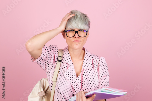 Stylish woman in dress and glasses on pink background with notebooks showing stressful worried touching head education concept