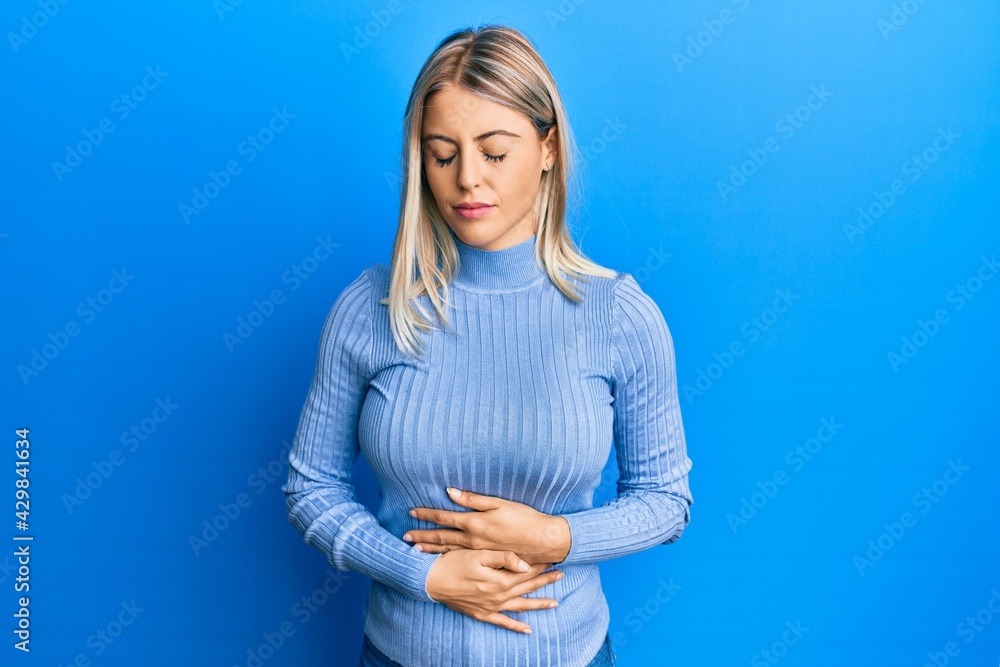 Beautiful blonde woman wearing casual clothes with hand on stomach because indigestion, painful illness feeling unwell. ache concept. Beautiful blonde woman wearing casual clothes with hand on stomach because indigestion, painful illness feeling unwell. ache concept.