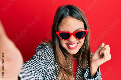 Young brunette woman taking a selfie photo wearing sunglasses screaming proud, celebrating victory and success very excited with raised arm © Krakenimages.com