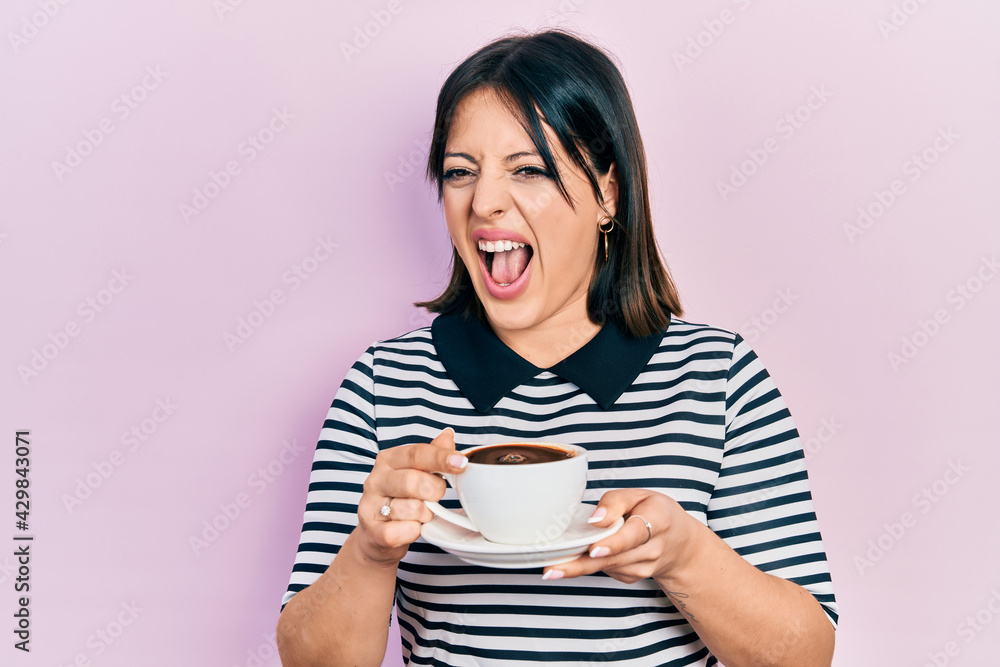 Young hispanic woman drinking a cup of coffee angry and mad screaming frustrated and furious, shouting with anger. rage and aggressive concept. Young hispanic woman drinking a cup of coffee angry and mad screaming frustrated and furious, shouting with anger. rage and aggressive concept.