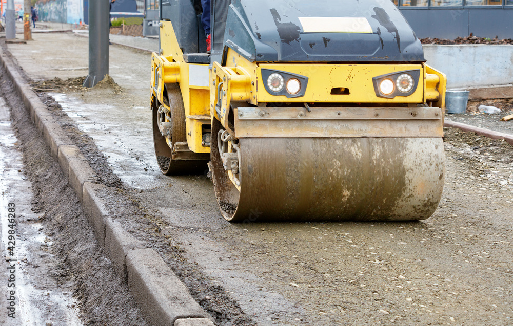 Frontal view of a road roller rolling the base of the sidewalk along a concrete curb near the carriageway.