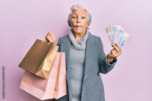 Senior grey-haired woman holding shopping bags and swedish krona banknotes making fish face with mouth and squinting eyes, crazy and comical.