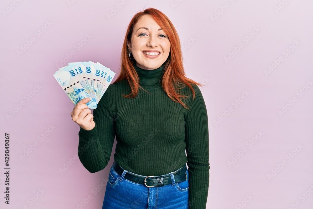 Beautiful redhead woman holding 20 hong kong dollars banknotes looking positive and happy standing and smiling with a confident smile showing teeth Beautiful redhead woman holding 20 hong kong dollars banknotes looking positive and happy standing and smiling with a confident smile showing teeth