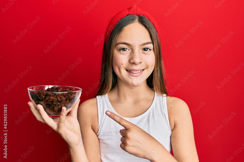 Beautiful brunette little girl holding bowl of raisins smiling happy pointing with hand and finger Beautiful brunette little girl holding bowl of raisins smiling happy pointing with hand and finger