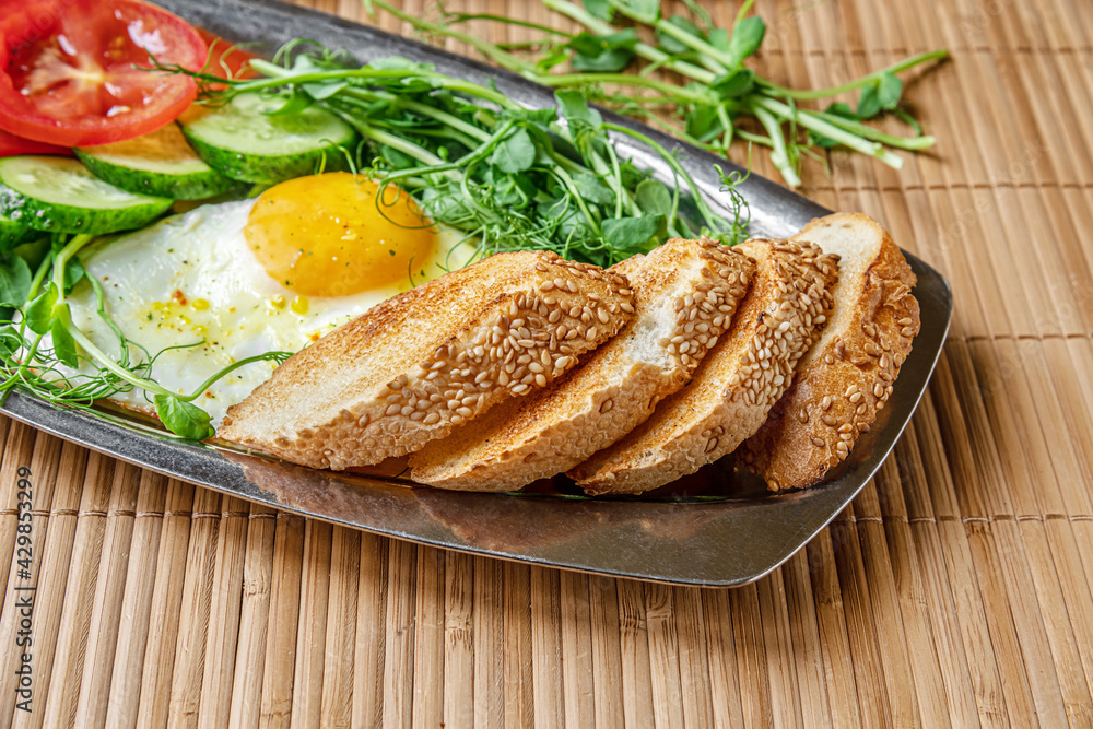 fried egg, toasted bread, cucumber and tomato slices and microgreens on a metal plate on bamboo straw surface, healthy breakfast concept fried egg, toasted bread, cucumber and tomato slices and microgreens on a metal plate on bamboo straw surface, healthy breakfast concept