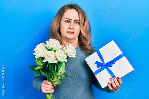 Middle age caucasian woman holding anniversary present and bouquet of flowers skeptic and nervous, frowning upset because of problem. negative person.