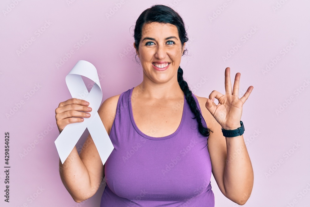 Plus size brunette woman holding white ribbon doing ok sign with fingers, smiling friendly gesturing excellent symbol Plus size brunette woman holding white ribbon doing ok sign with fingers, smiling friendly gesturing excellent symbol