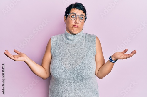 Plus size brunette woman wearing casual clothes and glasses clueless and confused expression with arms and hands raised. doubt concept.