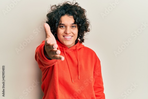 Young hispanic woman with curly hair wearing casual sweatshirt smiling friendly offering handshake as greeting and welcoming. successful business.