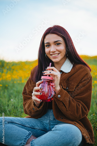 Smiling teenager girl drinking water in a red jar sitting on the grass of a hill. She is looking at camera. photo