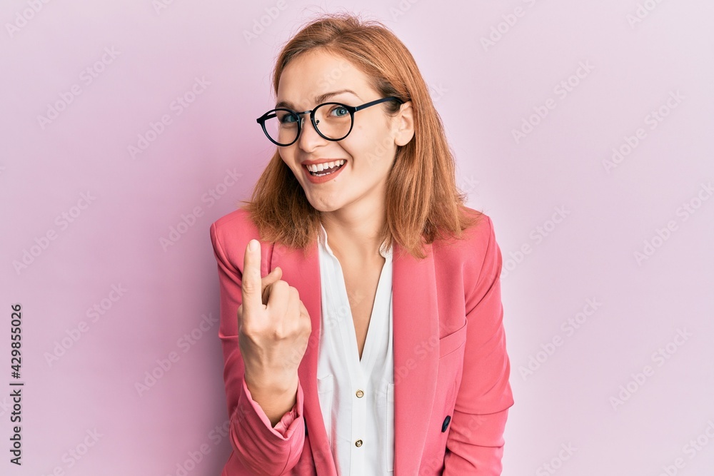 Young caucasian woman wearing business style and glasses beckoning come here gesture with hand inviting welcoming happy and smiling