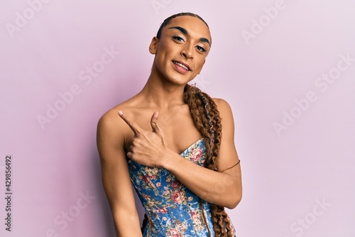 Hispanic man wearing make up and long hair wearing elegant corset cheerful with a smile of face pointing with hand and finger up to the side with happy and natural expression on face