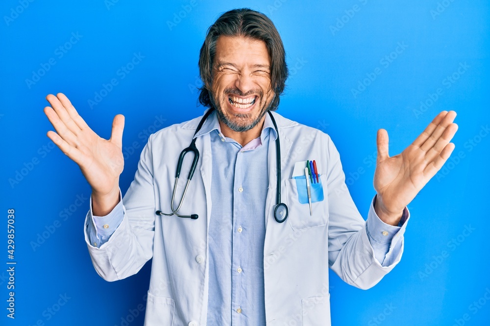 Middle age handsome man wearing doctor uniform and stethoscope celebrating mad and crazy for success with arms raised and closed eyes screaming excited. winner concept Middle age handsome man wearing doctor uniform and stethoscope celebrating mad and crazy for success with arms raised and closed eyes screaming excited. winner concept