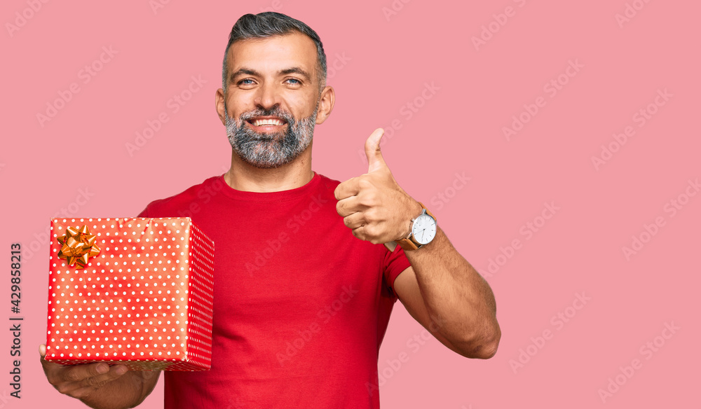 Middle age handsome man holding gift smiling happy and positive, thumb up doing excellent and approval sign