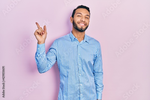 Hispanic man with beard wearing casual blue shirt with a big smile on face, pointing with hand and finger to the side looking at the camera.