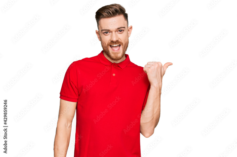Young redhead man wearing casual clothes smiling with happy face looking and pointing to the side with thumb up. Young redhead man wearing casual clothes smiling with happy face looking and pointing to the side with thumb up.