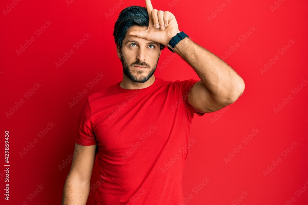 Fototapeta premium Young hispanic man wearing casual red t shirt making fun of people with fingers on forehead doing loser gesture mocking and insulting.